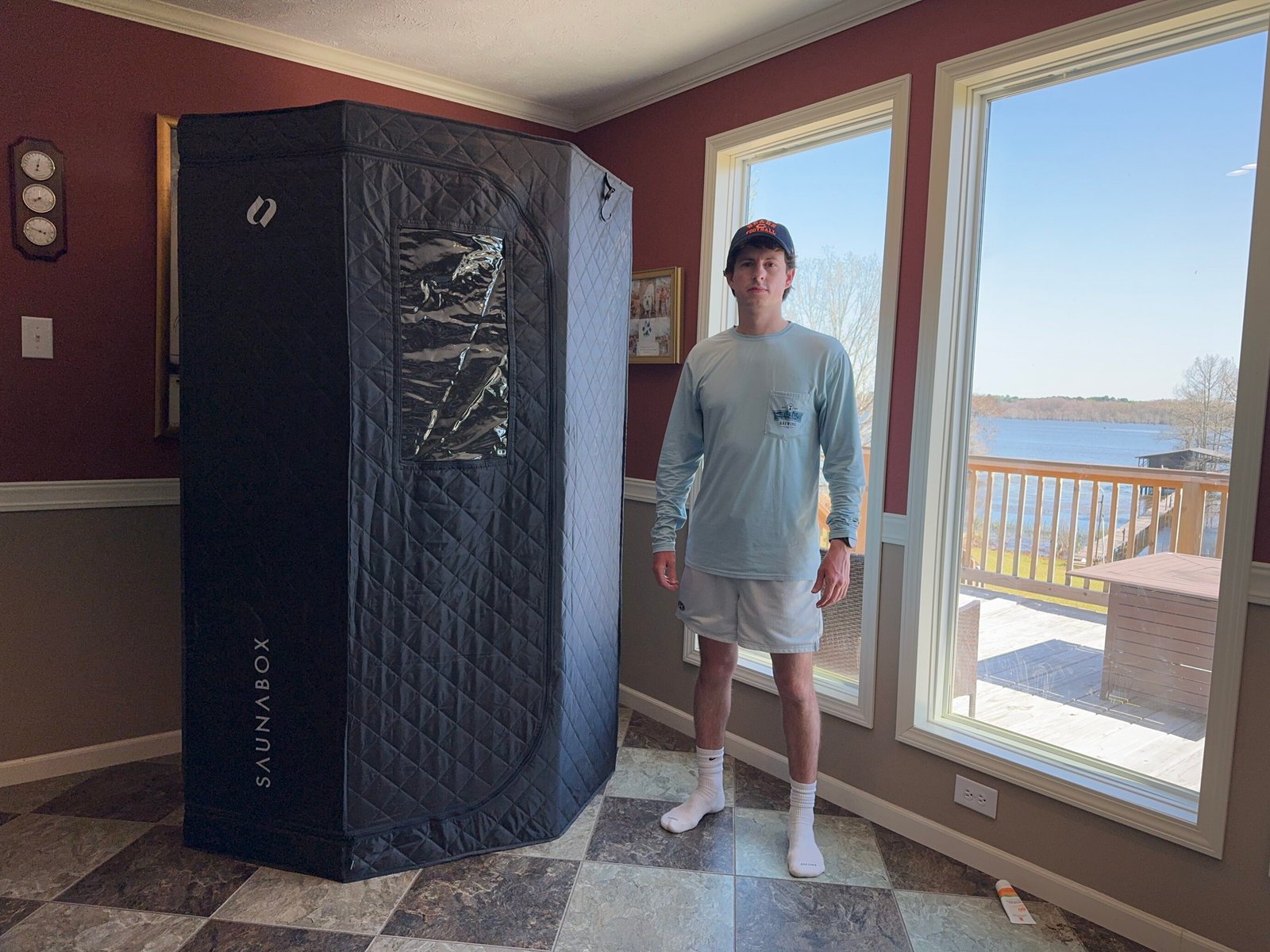 Bennett Carby standing beside a SaunaBox SmartSteam Pro portable sauna, positioned indoors, demonstrating the size and setup of the sauna system.
