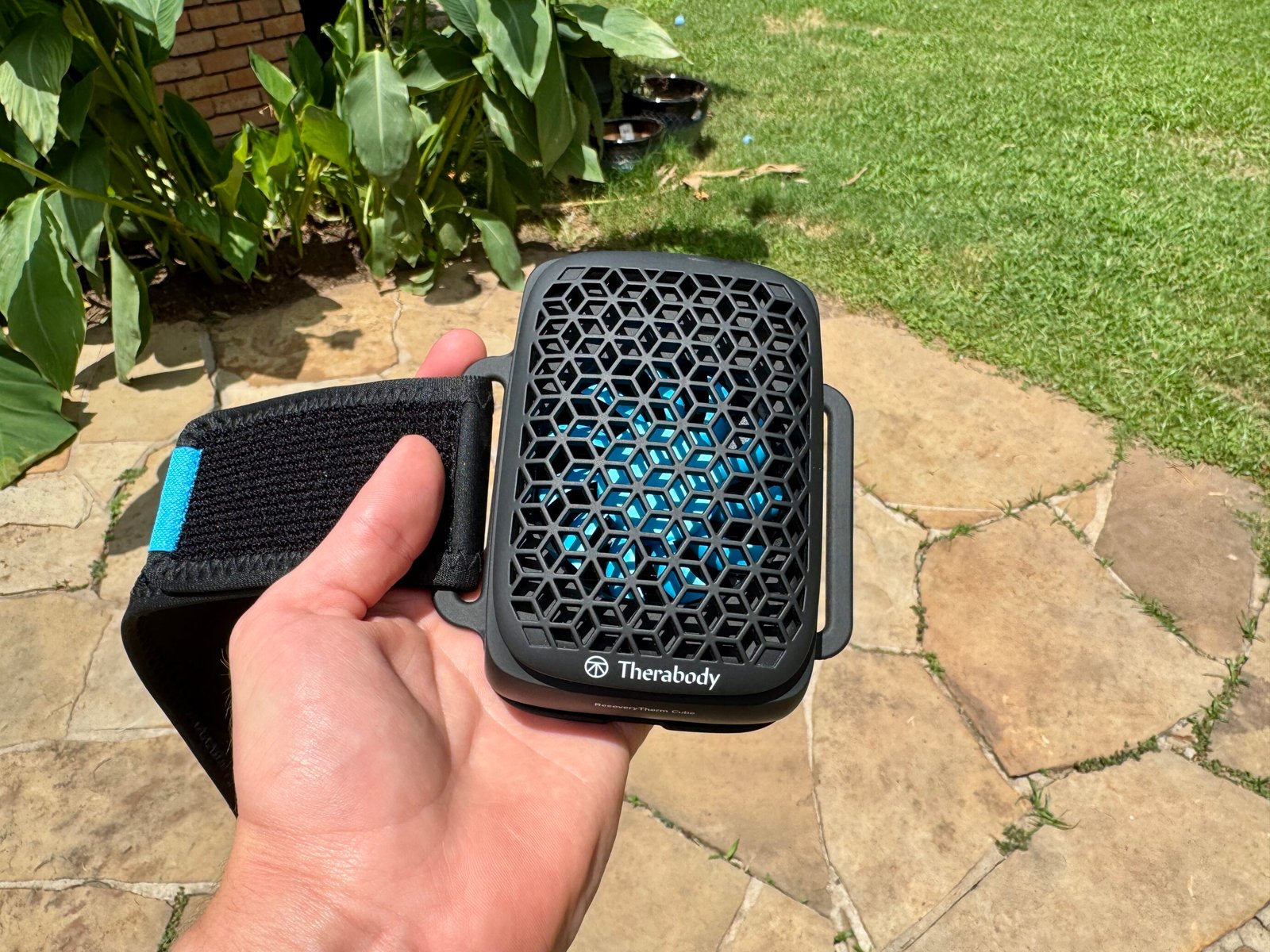 Therabody RecoveryTherm Cube held in hand showing the front hexagonal mesh grille with blue thermoelectric plate visible beneath, attached to a black neoprene velcro strap, photographed outdoors on a stone patio.