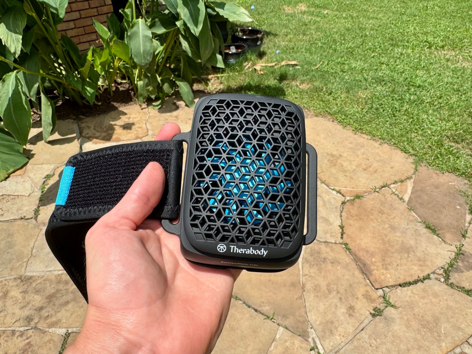 Therabody RecoveryTherm Cube held in hand showing the front hexagonal mesh grille with blue thermoelectric plate visible beneath, attached to a black neoprene velcro strap, photographed outdoors on a stone patio.