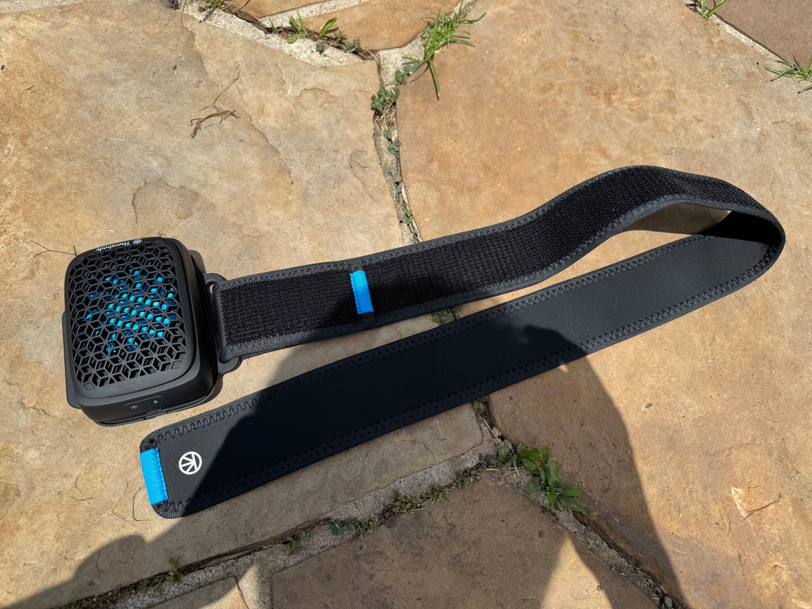 Therabody RecoveryTherm Cube with its full black neoprene velcro wrap strap laid flat on a stone surface, showing the device attached at one end and the Therabody logo tab at the other end.