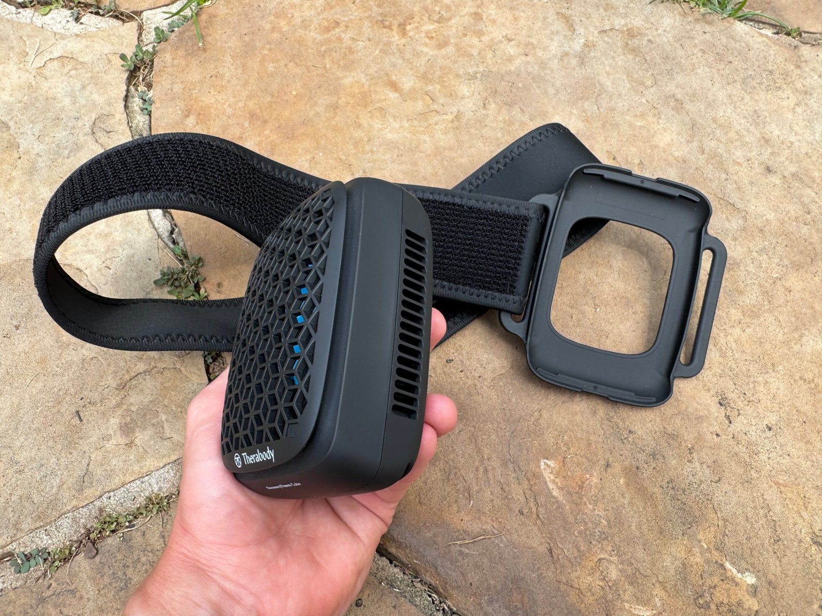 Therabody RecoveryTherm Cube removed from its plastic mounting bracket, held in hand alongside the detached strap and bracket assembly, showing the side vents.