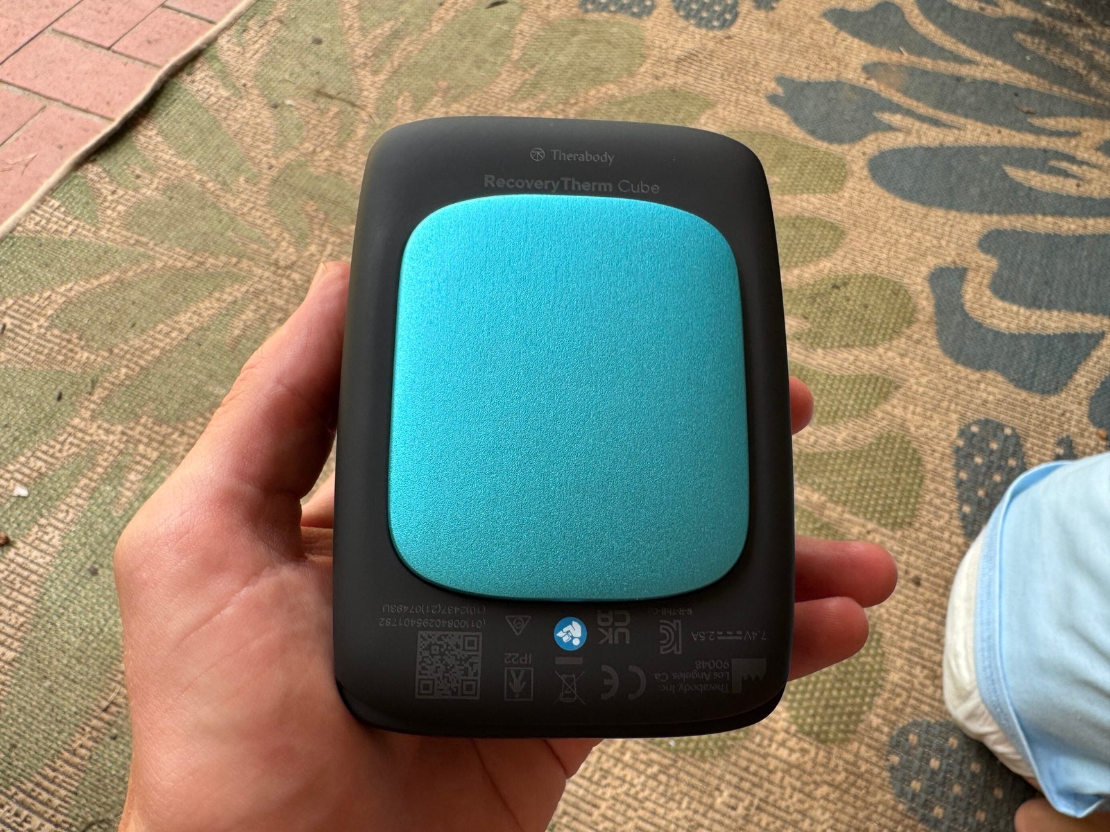 Back of the Therabody RecoveryTherm Cube showing the teal/cyan metallic thermoelectric contact plate, Therabody and RecoveryTherm Cube branding, and regulatory markings including IP22 rating and CE certification.